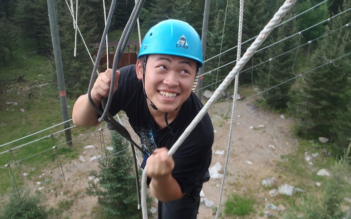 A person wearing safety gear and secured by ropes smiles as they navigate an obstacle of a high ropes course. 
